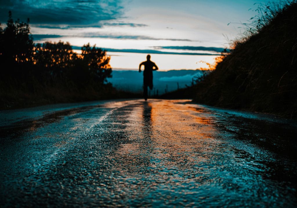 How to Stay Motivated During Long-Term Training Programs: Man running with sun set in background on a long road and trees at side